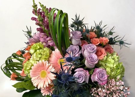 A lovely combination of pinks, purples, and greens arranged in a compact cylinder ensure this arrangement makes a lasting impression. Gerbera daises, spray roses, thistle, hydrangea, roses, and snapdragons are paired with unique greenery to give this arrangement its tropical and unique feel. 