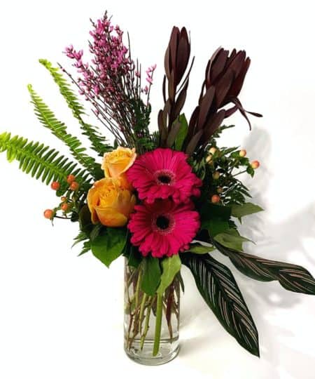 Bright gerbera daises, roses, and tropical foliage are paired together in this unique arrangement for a stunning display.